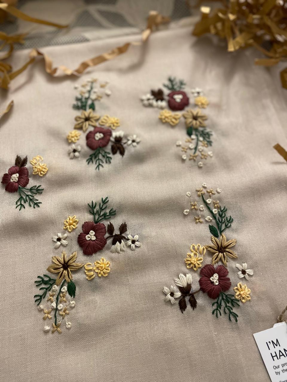 Détails des motifs floraux brodés à la main sur l'écharpe Lana Beige, fleurs bordeaux, jaunes et vertes sur tissu beige.