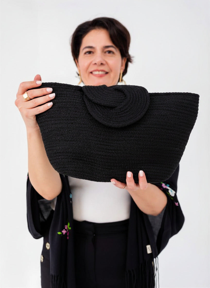 Gros plan sur le sac Nout noir pailleté, tenu à deux mains par un mannequin, montrant la texture du tressage et le détail de son rabat à fermoir graphique.