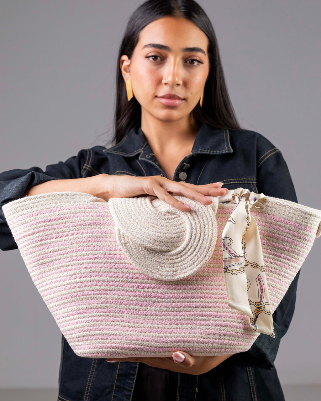 Gros plan sur le sac Héliopolis blanc à rayures roses, tenu à deux mains par un mannequin, montrant la texture du tressage et le détail du foulard en satin aux motifs graphiques.
